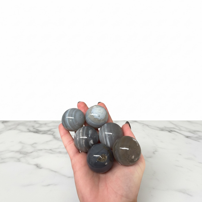 Kindred Crystal Collective — Agate Banded Sphere, four polished blue-grey banded agate spheres with concentric white banding, size reference held in open palm