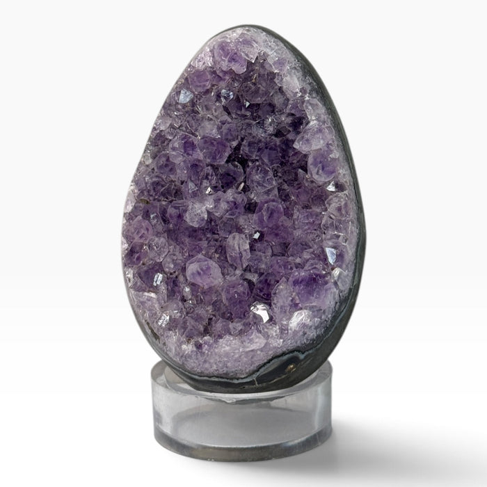 Kindred Crystal Collective — Druzy Amethyst Cluster Egg, egg-shaped stone with sparkling lilac and purple crystal points, a dark outer edge and a clear display stand.