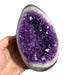 Kindred Crystal Collective — Amethyst Druzy Egg, hand-held open-face Amethyst druzy egg showing densely packed deep violet crystal points with white agate lining and natural basalt rind, size reference
