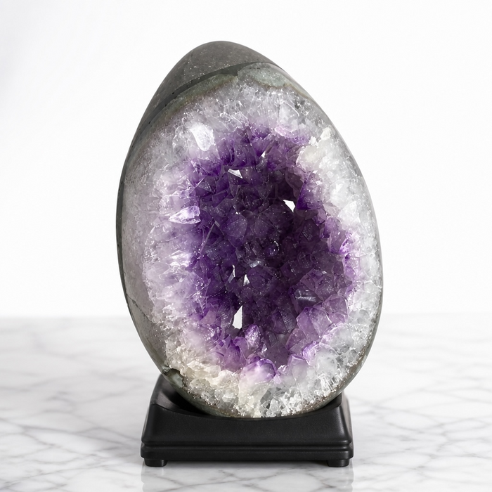 Kindred Crystal Collective — Amethyst Druzy Egg, polished dark grey Amethyst druzy egg on black wooden base showing pale lavender and violet crystal cavity, displayed on white marble surface