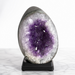 Kindred Crystal Collective — Amethyst Druzy Egg, polished dark grey Amethyst druzy egg on black wooden base showing pale lavender and violet crystal cavity, displayed on white marble surface