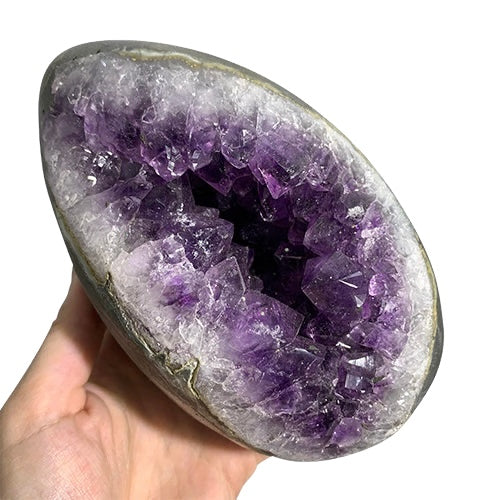 Kindred Crystal Collective — Amethyst Geode Egg, hand-held open Amethyst geode egg showing deep violet crystal interior with white agate lining and dark outer rind, size reference