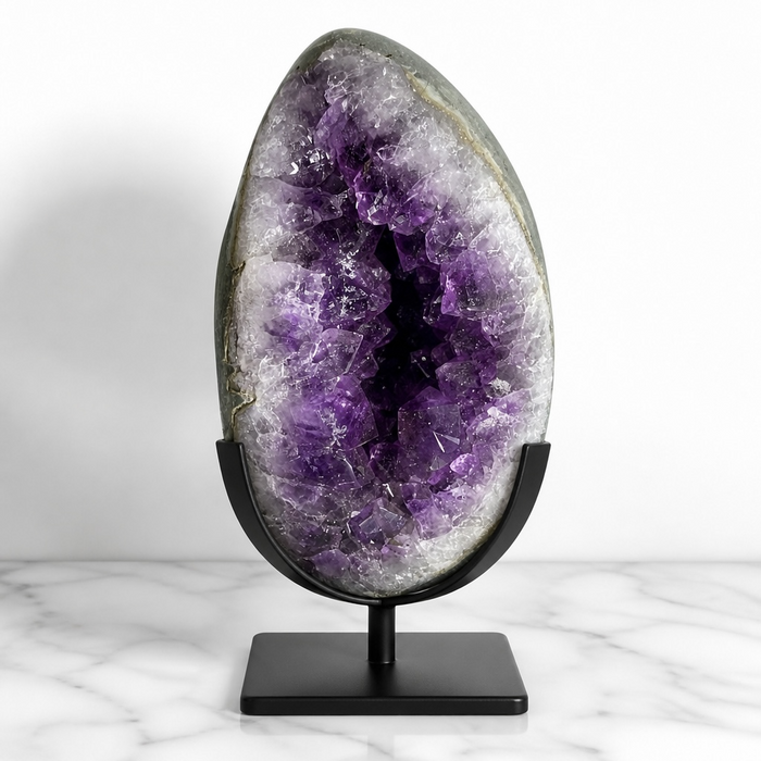 Kindred Crystal Collective — Amethyst Geode Egg, large open Amethyst geode egg on black metal stand showing deep violet crystal cavity and white agate lining, displayed on marble surface