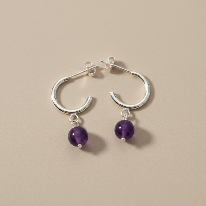 Kindred Crystal Collective - A pair of silver hoop earrings with dangling purple amethyst beads, displayed on a solid beige background.