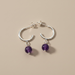 Kindred Crystal Collective - A pair of silver hoop earrings with dangling purple amethyst beads, displayed on a solid beige background.