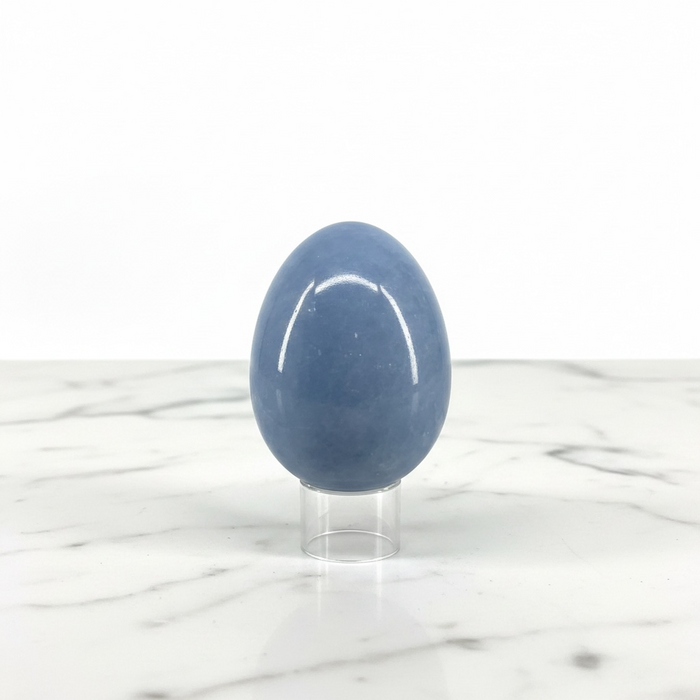 A polished blue stone egg displayed upright on a clear acrylic stand against a white marble surface and white background.