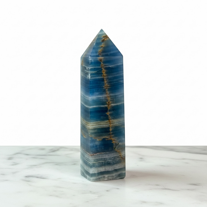 A polished blue onyx obelisk with natural gold veining, standing upright on a white marble surface against a white background.