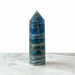 A polished blue onyx obelisk with natural gold veining, standing upright on a white marble surface against a white background.