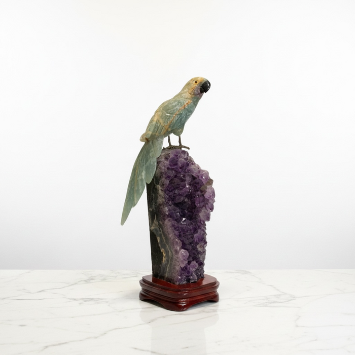Kindred Crystal Collective — A carved parrot figurine with iridescent green and gold feathers perched on a purple amethyst geode cluster mounted on a polished reddish-brown wooden base, set against a white studio background.