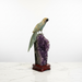 Kindred Crystal Collective — A carved parrot figurine with iridescent green and gold feathers perched on a purple amethyst geode cluster mounted on a polished reddish-brown wooden base, set against a white studio background.