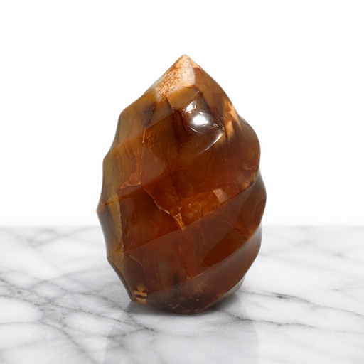 Kindred Crystal Collective — Carnelian Flame, hand-carved deep amber-brown Carnelian flame with faceted high-gloss polish and natural internal banding, displayed on white marble surface