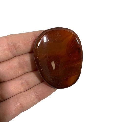Kindred Crystal Collective — Carnelian Worry Stone, polished oval reddish-brown carnelian with subtle colour variation held between fingers against a white background.