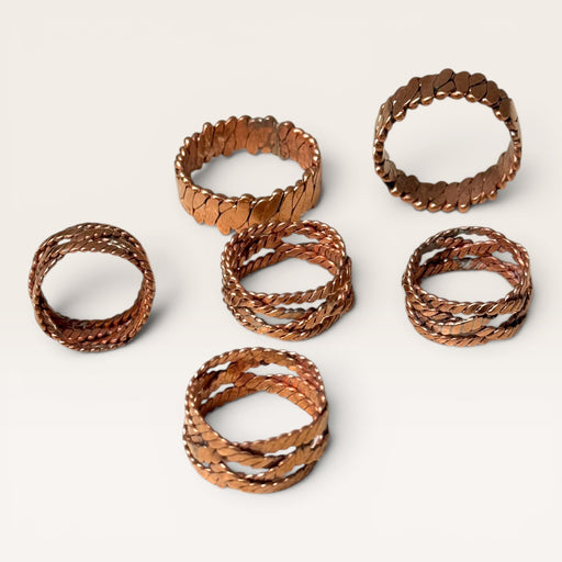 Kindred Crystal Collective - Six copper-toned metal rings with textured, hammered, and twisted finishes arranged on a light beige background.