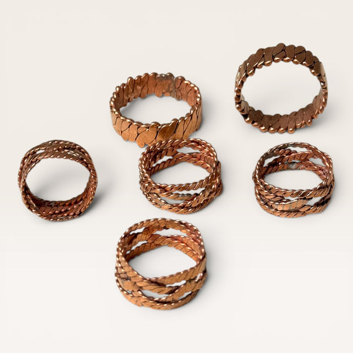 Kindred Crystal Collective - Six copper-toned metal rings with textured, hammered, and twisted finishes arranged on a light beige background.