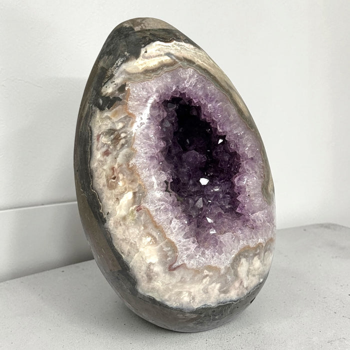 Kindred Crystal Collective — Druzy Amethyst Egg, sparkling purple crystal cavity framed by polished cream, taupe and grey banding, on a light surface.