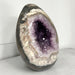 Kindred Crystal Collective — Druzy Amethyst Egg, sparkling purple crystal cavity framed by polished cream, taupe and grey banding, on a light surface.