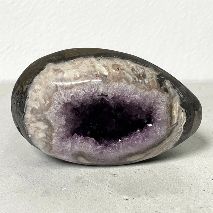 Kindred Crystal Collective — Druzy Amethyst Egg, sparkling purple crystal cavity framed by polished cream, taupe and grey banding, on a light surface.