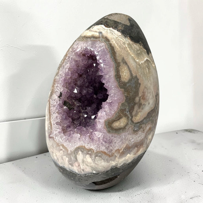 Kindred Crystal Collective — Druzy Amethyst Egg, sparkling purple crystal cavity framed by polished cream, taupe and grey banding, on a light surface.