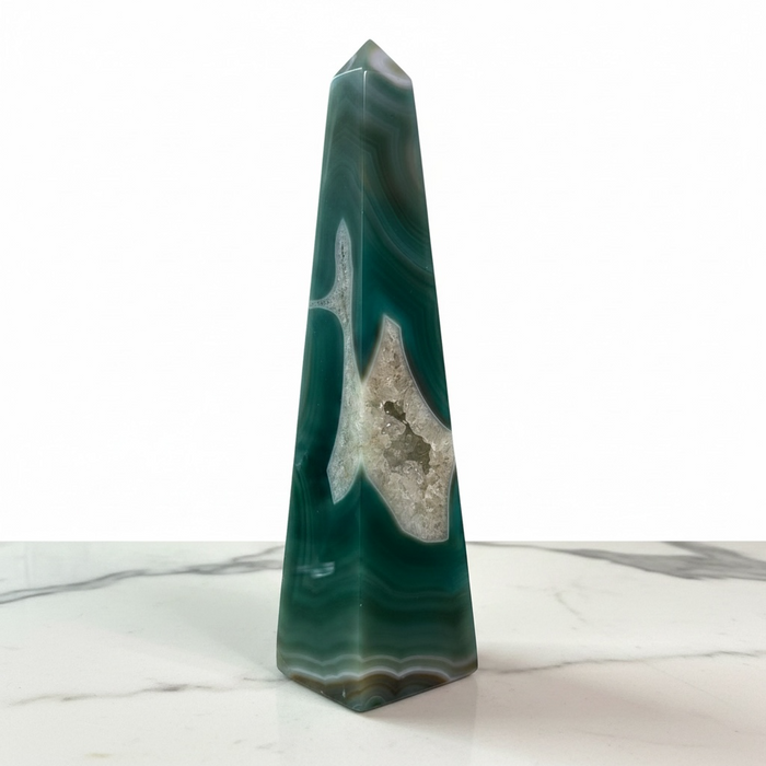 Kindred Crystal Collective - A polished natural agate crystal obelisk with green, teal, and white banded patterns and a prominent white quartz vein, standing on a white marble surface against a white background.