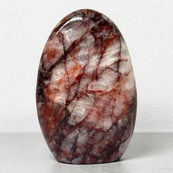 Kindred Crystal Collective — Hematoid Quartz Freeform, polished upright quartz with burgundy and rust-red inclusions through cloudy white stone, on a light surface.