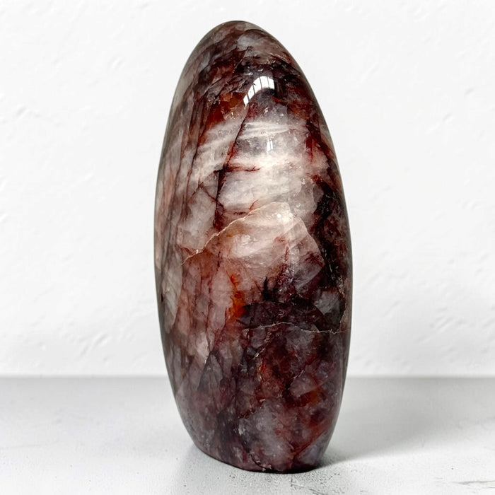 Kindred Crystal Collective — Hematoid Quartz Freeform, polished upright quartz with burgundy and rust-red inclusions through cloudy white stone, on a light surface.