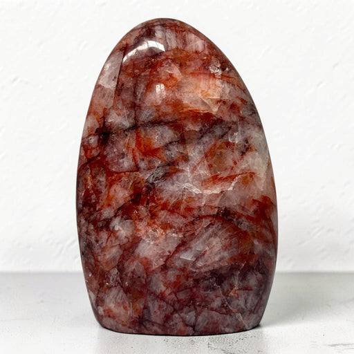 Kindred Crystal Collective — Hematoid Quartz Freeform, polished rounded quartz display piece with copper-red inclusions, burgundy seams and cloudy white patches, on a light surface.