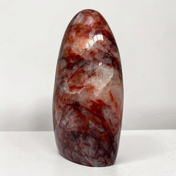 Kindred Crystal Collective — Hematoid Quartz Freeform, polished rounded quartz display piece with copper-red inclusions, burgundy seams and cloudy white patches, on a light surface.
