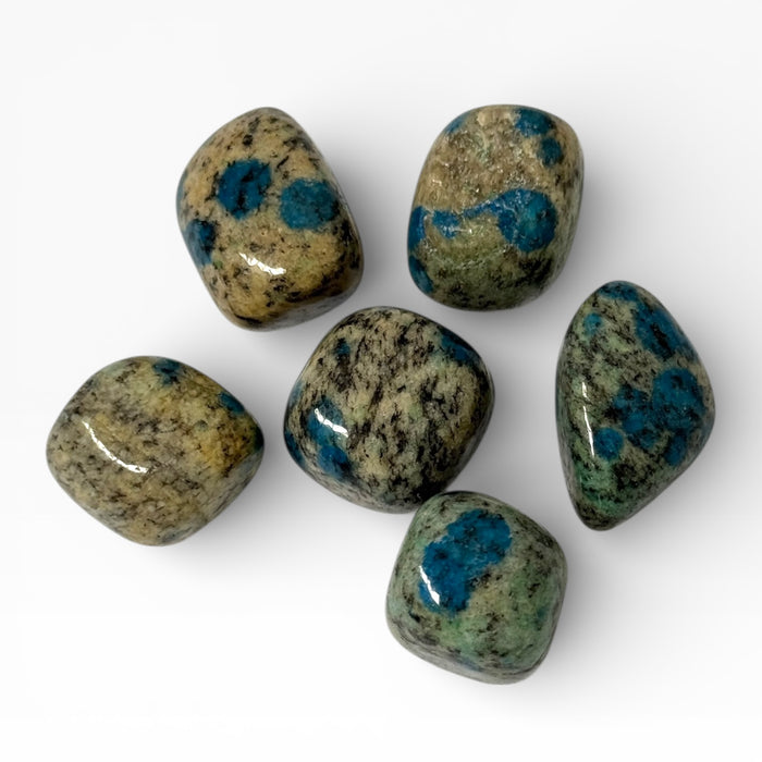 Kindred Crystal Collective — K2 Stone Tumbled Stones, six polished K2 granite tumbles showing cream-beige speckled granite matrix with vivid teal-blue Azurite spots, white background