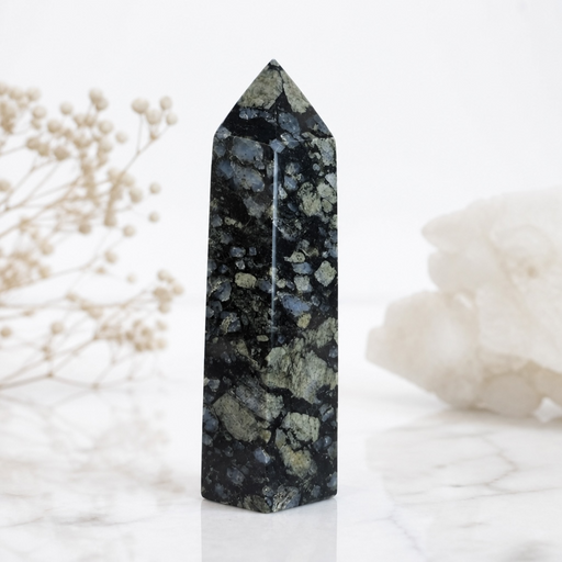 Kindred Crystal Collective — Llanite Generator Grey Matrix, polished dark grey Llanite crystal tower with pale blue-grey feldspar inclusions, displayed on white marble with dried floral styling