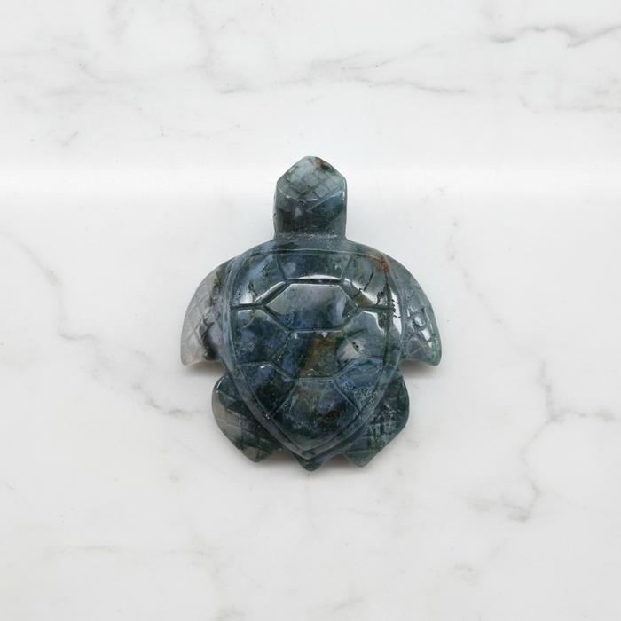 Kindred Crystal Collective - Hand-carved Moss Agate turtle figurine held in a hand, showing deep grey-green stone with detailed shell and flipper carving.