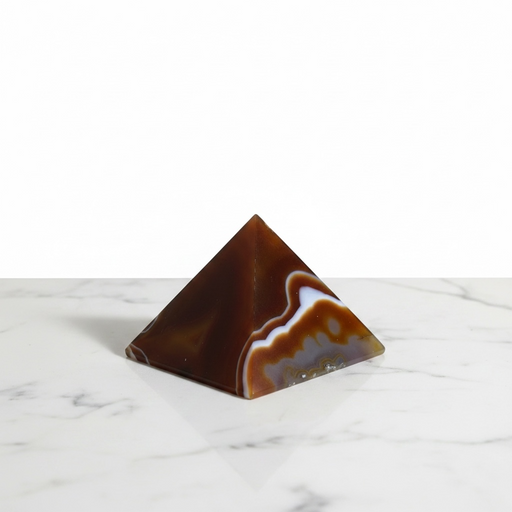 Kindred Crystal Collective - A polished pyramid agate crystal pyramid with natural banding in dark brown, amber, and white, displayed on a white marble surface against a white background.