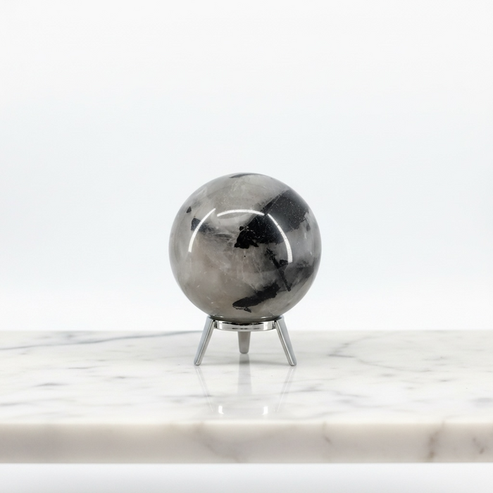 A Quartz Tourmaline Sphere crystal on a small silver metal stand, displayed on a white marble surface against a clean white background.