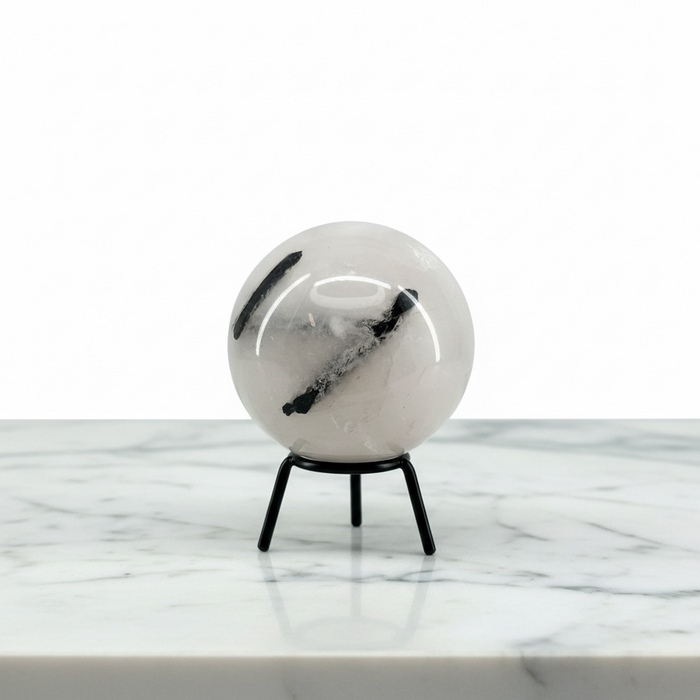 A Quartz Tourmaline Sphere with black inclusions, displayed on a small black metal stand on a white marble surface against a white background.
