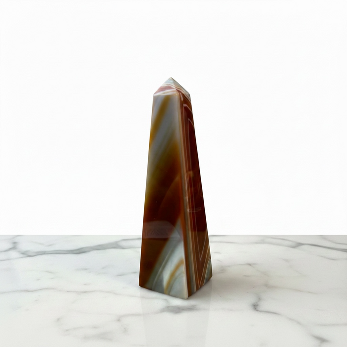 Kindred Crystal Collective - A polished natural agate crystal obelisk with swirling bands of brown, reddish-brown, cream, and white, standing upright on a white marble surface against a white background.