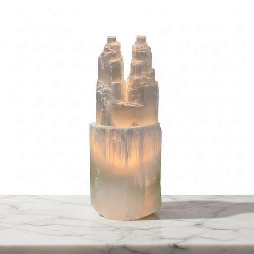 Kindred Crystal Collective - A Selenite Twin Tower Lamp - 30cm lamp with a cylindrical base sits on a white marble surface against a white background.