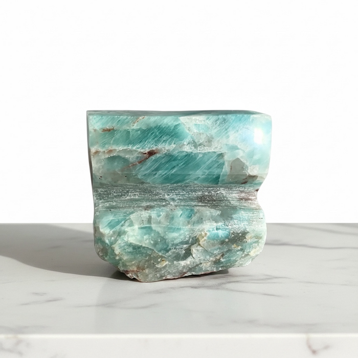 Kindred Crystal Collective - A polished raw amazonite crystal sculpture with natural wave-like patterns, displayed on a white marble surface against a white background.