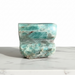 Kindred Crystal Collective - A polished raw amazonite crystal sculpture with natural wave-like patterns, displayed on a white marble surface against a white background.