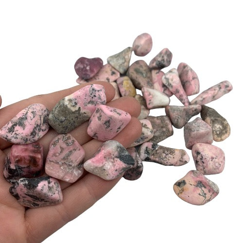 Kindred Crystal Collective — Rhodonite Tumbled 1.5–3cm, hand holding a pile of polished dusty pink and rose stones with dark grey-black manganese veining