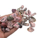 Kindred Crystal Collective — Rhodonite Tumbled 1.5–3cm, hand holding a pile of polished dusty pink and rose stones with dark grey-black manganese veining